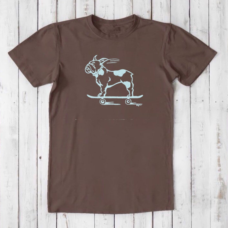 May include: A brown t-shirt with a light blue graphic of a French bulldog skateboarding. The bulldog is wearing a collar and has its tongue sticking out.
