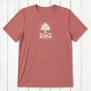 May include: A dusty rose short-sleeve t-shirt featuring a cream-colored graphic of a tree with visible roots inside a rectangular frame. Above the tree is a small crescent moon. The text "grow more" is printed in lowercase below the tree.