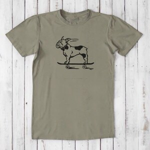 May include: A khaki green t-shirt with a black and white illustration of a bulldog skateboarding. The bulldog is wearing a collar and has a tongue sticking out. The skateboard has wheels and the text "Bugger" is printed below the skateboard.