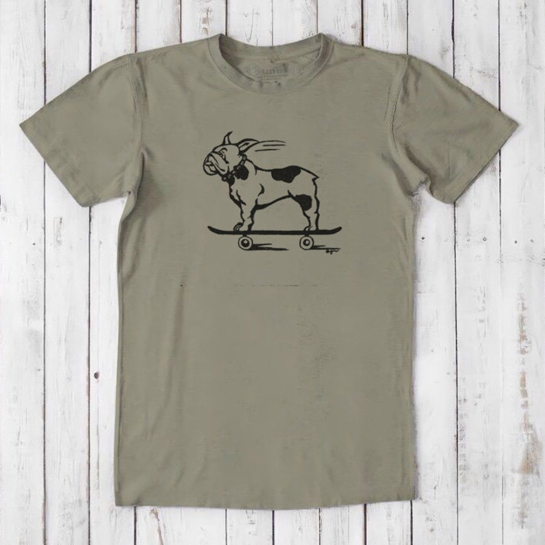 May include: A khaki green t-shirt with a black and white illustration of a bulldog skateboarding. The bulldog is wearing a collar and has a tongue sticking out. The skateboard has wheels and the text "Bugger" is printed below the skateboard.