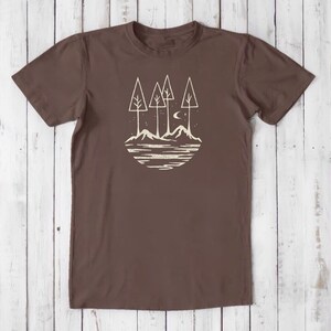 May include: A brown t-shirt with a white graphic of a stylized landscape with trees, mountains, a crescent moon, and stars.