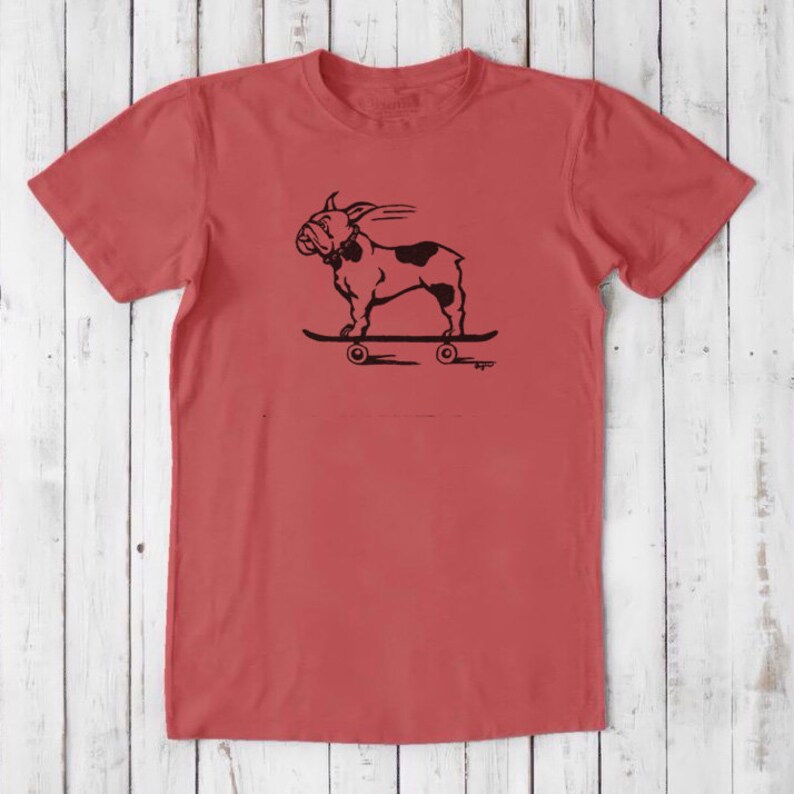 May include: A red t-shirt with a black graphic of a bulldog skateboarding. The bulldog is wearing a collar and has a tongue sticking out. The skateboard has four wheels.
