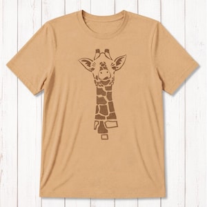 May include: A tan short-sleeved t-shirt featuring a centered, dark brown graphic print of a giraffe head and neck. The giraffe design is composed of distinct, blocky, and irregular shapes that form the animal's facial features and spotted pattern.