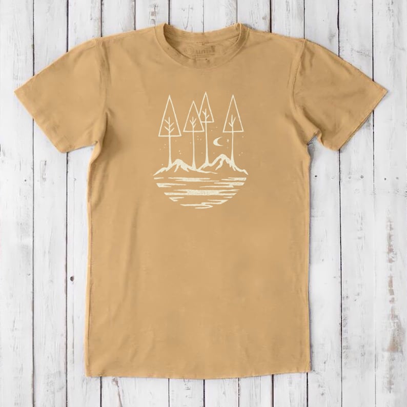 May include: A tan t-shirt with a white graphic of a moon, three trees, and mountains reflected in a lake.