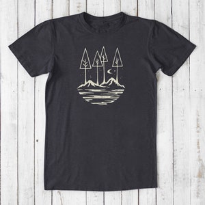 May include: A dark gray t-shirt with a white graphic design of a stylized forest scene with three trees, mountains, a crescent moon, and stars. The design is in a circle.