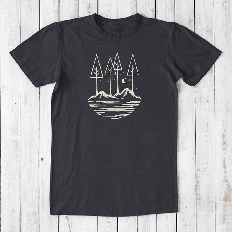 May include: A dark gray t-shirt with a white graphic design of a stylized forest scene with three trees, mountains, a crescent moon, and stars. The design is in a circle.