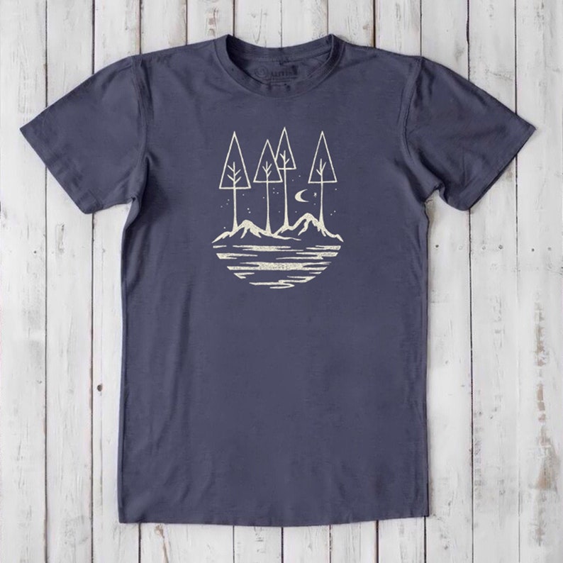 May include: A navy blue t-shirt with a white graphic design of a stylized landscape with four trees, mountains, a crescent moon, and a lake.