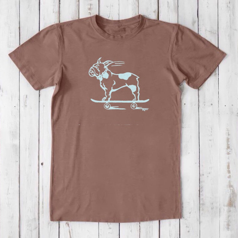 May include: A brown t-shirt with a light blue graphic of a French bulldog skateboarding. The graphic is on the front of the shirt.