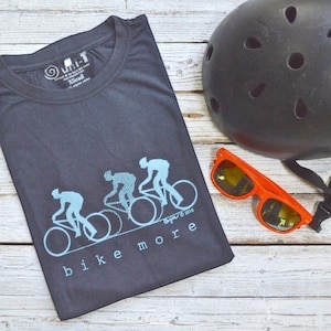 May include: A dark gray short-sleeve t-shirt featuring a light blue graphic of three cyclists and the text "bike more". The shirt is displayed alongside a black bicycle helmet and orange-framed sunglasses with dark lenses.