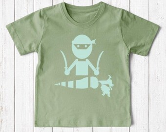 May include: A sage green short-sleeve t-shirt featuring a light mint green graphic print of a stylized ninja figure holding two swords, positioned above a horizontal carrot shape with a leafy top on the center of the chest.