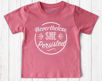 May include: A short-sleeved crew neck t-shirt in a dusty rose pink color. The center of the shirt features a white circular graphic containing the text "Nevertheless SHE Persisted" in a stylized white font with decorative lines.