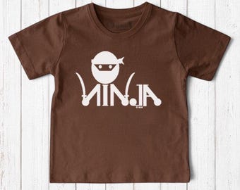May include: A brown short-sleeved t-shirt featuring a white graphic of a stylized ninja head with a mask and the word NINJA written in a mirrored, decorative font with small sword icons integrated into the lettering and a 2018 copyright.