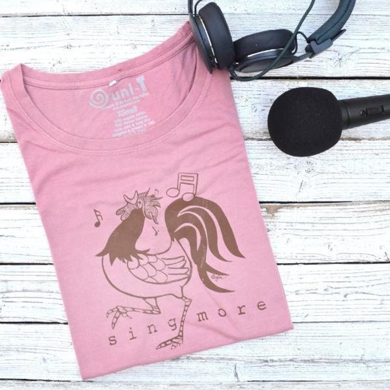 May include: A pink t-shirt with a brown rooster illustration and the words 'sing more' printed on the front. The rooster is wearing headphones and has a musical note above its head.