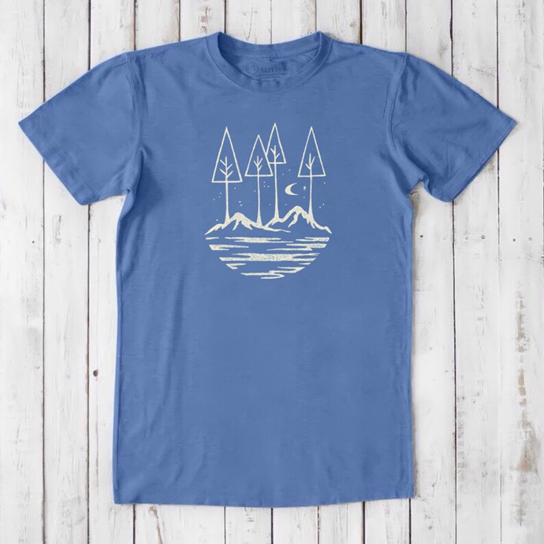 May include: A blue t-shirt with a white graphic of a moon, stars, and three trees on a mountain landscape.