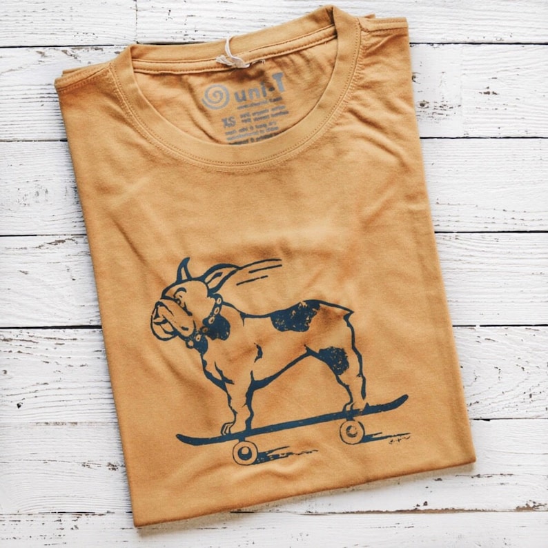 May include: A mustard yellow t-shirt with a blue graphic of a bulldog skateboarding. The t-shirt has a small tag on the front that says 'unit' and 'XS'.