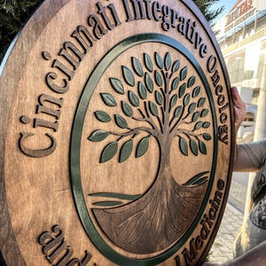 Puede incluir: Un letrero redondo de madera con las palabras "Cincinnati Integrative Oncology and Functional Medicine" talladas alrededor del borde. Un diseño de árbol en el centro, con detalles verdes.