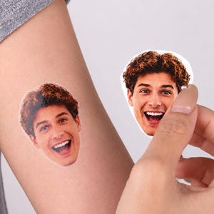 May include: A temporary tattoo featuring a photographic portrait of a smiling person with brown curly hair. The tattoo is applied to skin, and an identical adhesive sticker version of the same portrait is held up nearby for comparison.
