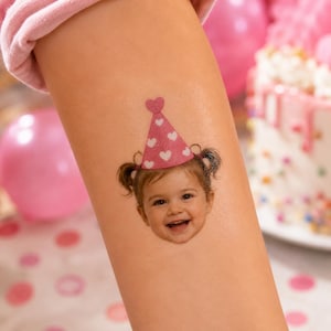 Può includere: Un tatuaggio temporaneo raffigurante il volto di un bambino sorridente con capelli castani raccolti in due codini. Il bambino indossa un cappellino da festa rosa decorato con un motivo di piccoli cuori bianchi e un topper a forma di cuore.