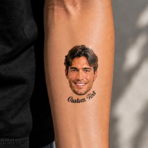 Op de afbeelding: Een tijdelijke tatoeage op de huid met een realistisch kleurenportret van een man met donkerbruin haar en een lichte baard, die lacht met zichtbare tanden. Onder het portret staan de woorden Custom Text in een zwart cursief lettertype.