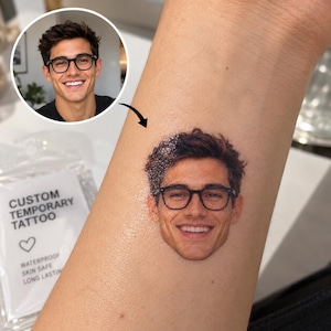 Könnte beinhalten: Ein auf die Haut aufgetragenes, individuelles temporäres Tattoo mit einem realistischen Farbporträt einer lächelnden Person mit dunklem Haar und schwarzer Brille. Daneben liegt eine Klarsichtverpackung mit der Aufschrift CUSTOM TEMPORARY TATTOO, WATERPROOF, SKIN SAFE.