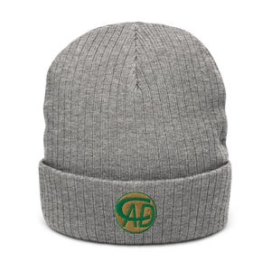 Puede incluir: Un gorro de punto acanalado gris con un borde doblado. El gorro presenta un emblema bordado con las letras "AD" en verde, dentro de un círculo dorado. El gorro está hecho de un material suave y texturizado.