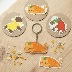 May include: A selection of stickers and a keychain with cartoon animal designs. The designs include a cat, a dog in a banana costume, and a bunny in a strawberry costume. The keychain and stickers are orange and white. The text "Handmade in the UK - Custom Options Available" is at the bottom.