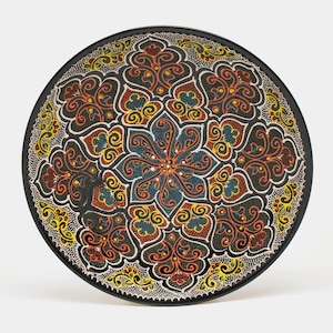 May include: A round, decorative plate with a black rim and a detailed, colorful floral pattern. The design features a central flower with blue petals, surrounded by red, yellow, and orange scrollwork on a black background. The plate's edge has a dotted pattern.