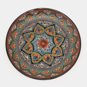 May include: A decorative ceramic plate with a black rim and a detailed floral pattern. The plate's design includes a central starburst in orange, surrounded by symmetrical floral and geometric elements in blue, yellow, green, and red. The plate is round.