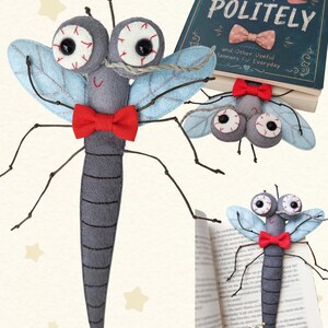 Könnte beinhalten: Ein graues Libellen-Stofftier aus Filz mit großen weißen Augen mit roten Adern, hellblauen Flügeln mit gestickten Details und einer roten Fliege. Das Spielzeug wird neben einem Buch mit dem Titel HOW TO BITE POLITELY und offenen Buchseiten gezeigt.