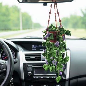 May include: A crocheted hanging plant in a car. The plant pot is brown, and the leaves are green with purple flowers. The plant hangs from the rearview mirror. The car's dashboard and steering wheel are visible in the foreground.