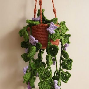 Puede incluir: Una planta colgante de ganchillo en una maceta color terracota con hojas de hiedra verdes y pequeñas flores moradas. El texto en la parte superior dice: Make Your Own Cozy Ivy Crochet Pattern PDF. La planta cuelga de tres cordones de ganchillo.