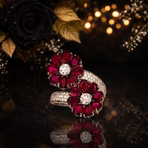 Può includere: Un anello a forma di fiore con pietre preziose rosse e un diamante centrale. La fascia dell'anello è incastonata con piccoli diamanti, creando un effetto scintillante. Il design è ambientato su uno sfondo scuro con luci sfocate.