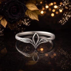 May include: A silver-toned ring featuring a central pointed design with engraved leaf-like patterns on each side. The band has a smooth, polished finish and a symmetrical shape that tapers toward the center, creating a distinct crown-like appearance.