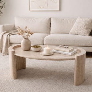 Könnte beinhalten: Ein Wohnzimmer in neutralen Tönen mit einem beigen Sofa, einem hellen Teppich und einem ovalen Couchtisch. Auf dem Tisch stehen eine Vase mit Trockenblumen, eine Kerze, Bücher und eine Schale. Eine dekorative Decke liegt auf dem Sofa.
