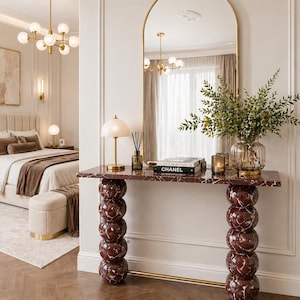 May include: An elegant bedroom scene with a marble console table. The table has stacked spherical legs and supports a vase of greenery, candles, and books. A large arched mirror reflects the room, including a bed with a tufted headboard.