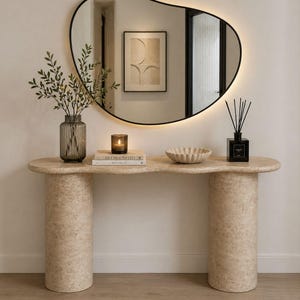 May include: A beige stone console table with two cylindrical supports. The table displays a vase of foliage, a candle, books, a decorative bowl, and a black diffuser. A large, black-framed mirror is above.