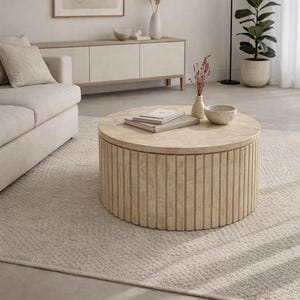 Puede incluir: Una mesa de centro redonda de madera clara con detalles de ranuras verticales. La mesa tiene libros y un cuenco pequeño en la parte superior. Está sobre una alfombra clara en un salón.
