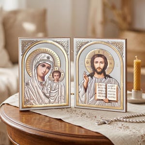 Puede incluir: Dos iconos religiosos con detalles plateados y dorados. Uno representa a María y Jesús, el otro a Jesús sosteniendo un libro. Ambos tienen halos dorados y están en marcos ornamentados. Los iconos están sobre una mesa de madera.