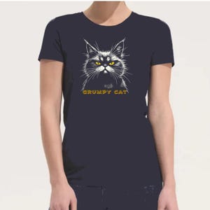 Op de afbeelding: Marineblauwe t-shirt met een illustratie van een chagrijnige kat met gele ogen en de tekst "GRUMPY CAT" in goud. De kat is gedetailleerd afgebeeld, wat een contrast creëert. Een casual grafisch T-shirt.