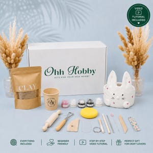 May include: A craft kit box labeled Ohh Hobby containing a brown clay pouch, a paper cup, five small paint pots, a yellow sponge, and various wooden and metal sculpting tools. A white ceramic rabbit planter with pink hearts sits nearby.