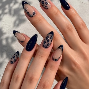 May include: Almond-shaped nails featuring a combination of solid dark navy blue polish and a nude base with dark navy blue leopard print patterns. Some nails display a French tip style with thin gold metallic lines separating the colors.