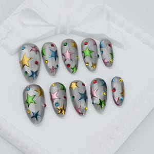 Peut inclure: Un ensemble de dix faux ongles en forme d'amande avec une base grise translucide. Chaque ongle présente une variété d'autocollants en forme d'étoiles métalliques dorées, roses, bleues et vertes, agrémentés de petits strass ronds colorés.