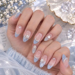Op de afbeelding: Ovaalvormige nagels met een lichtblauwe French manicure op een transparante basis. De nagels zijn versierd met witte parelaccenten, kleine zilveren strass-steentjes en verhoogde stervormige decoraties. Sommige nagels hebben een subtiel wit ruitpatroon.