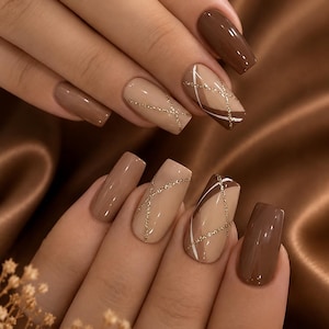 May include: Square-shaped nails featuring a manicure with alternating solid dark brown and light beige polish. The beige nails are decorated with thin, intersecting lines of gold glitter and white paint, creating a geometric pattern across the surface.