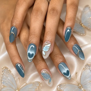 May include: Almond-shaped nails featuring a glossy slate blue cat-eye polish with metallic gold line accents. Two nails display a translucent base with a delicate gold-outlined blue butterfly wing design, creating a cohesive and detailed manicure.