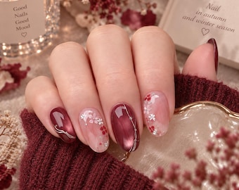May include: Manicured fingernails featuring a mix of deep burgundy glossy polish and translucent light pink polish with delicate silver and red floral accents. The nails are decorated with thin silver lines and small floral motifs, creating a sophisticated look.