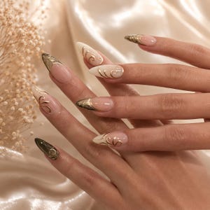 May include: Long almond-shaped nails featuring a French manicure style with a nude base and white or dark tips. Each nail is decorated with intricate gold metallic swirl patterns that extend from the tips toward the center of the nail bed.