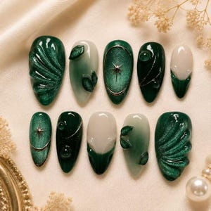 Könnte beinhalten: Ein Set aus zehn künstlichen Fingernägeln in den Farben Waldgrün und Creme. Die Designs umfassen strukturierte Wellenmuster, kleine grüne Blattakzente, goldene Sternanhänger, feine metallische Goldlinien und glatte, glänzende Oberflächen.