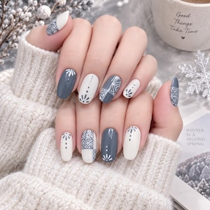 Op de afbeelding: Gemanicureerde vingernagels met een afwisselend kleurenschema van effen wit en leigrijs. De nagels zijn versierd met ingewikkelde witte lijntekeningen, waaronder geometrische patronen, kleine stippen en bloemachtige motieven op bepaalde nagels.
