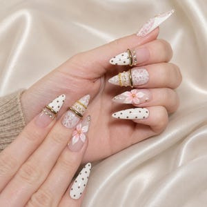 Puede incluir: Uñas postizas en forma de estilete con bases blancas y color carne, decoradas con lunares dorados, patrones de encaje blanco y flores rosas pintadas a mano. Varias uñas incluyen anillos dorados delgados y pequeños cristales transparentes.